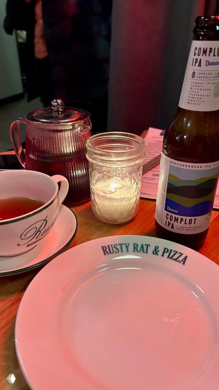 Стол с тарелкой Rusty Rat & Pizza, бутылкой IPA, чайником и зажжённой стеклянной свечой — сочетание пиццы и напитков в заведении.