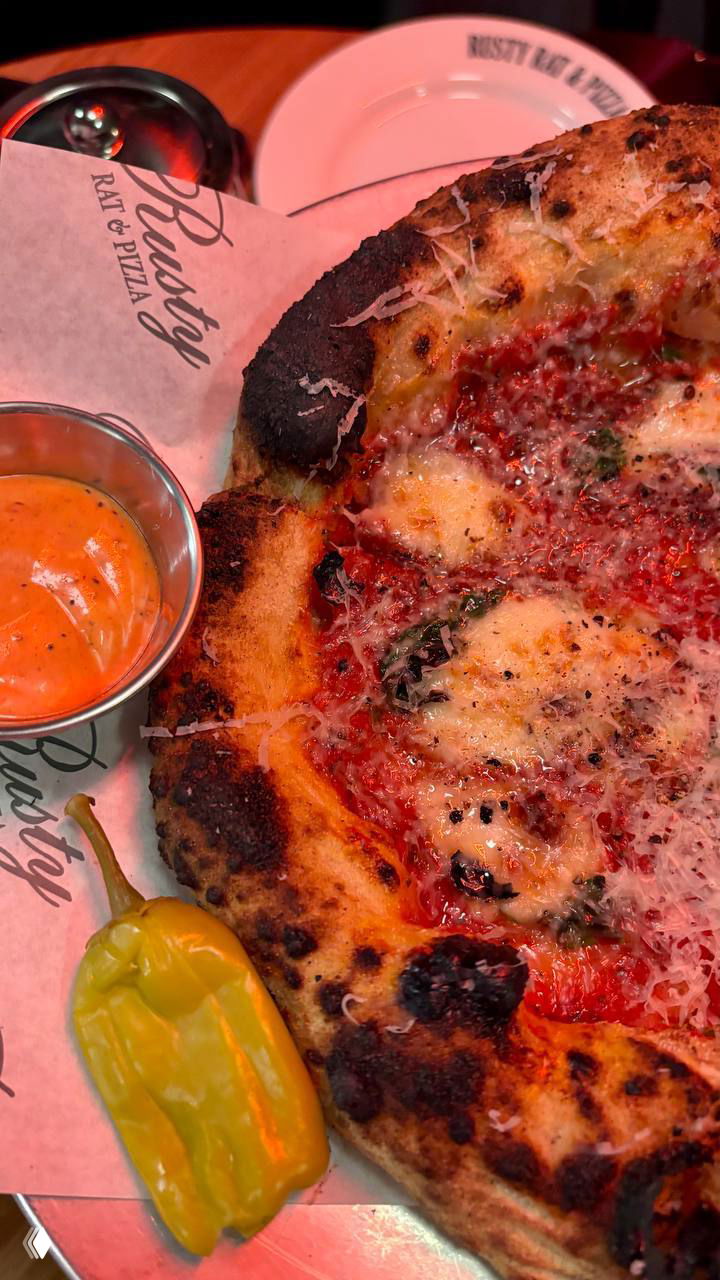 Крупный план пиццы с подпалённым бортиком и соусом в металлической чашке; рядом целый желтый острый перчик как закуска.