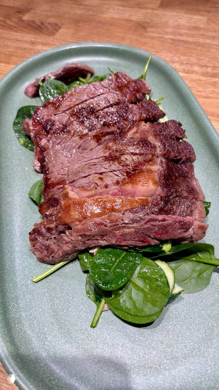 Крупный кусок прожаренного стейка на зелёной тарелке с листьями шпината на деревянном столе — подача в One Chef Studio.