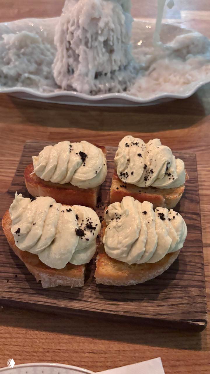 Небольшие брускетты с пышной ореховой или сливочной шапкой на деревянной доске, на фоне ещё одного десерта — подача закуски и сладкого.