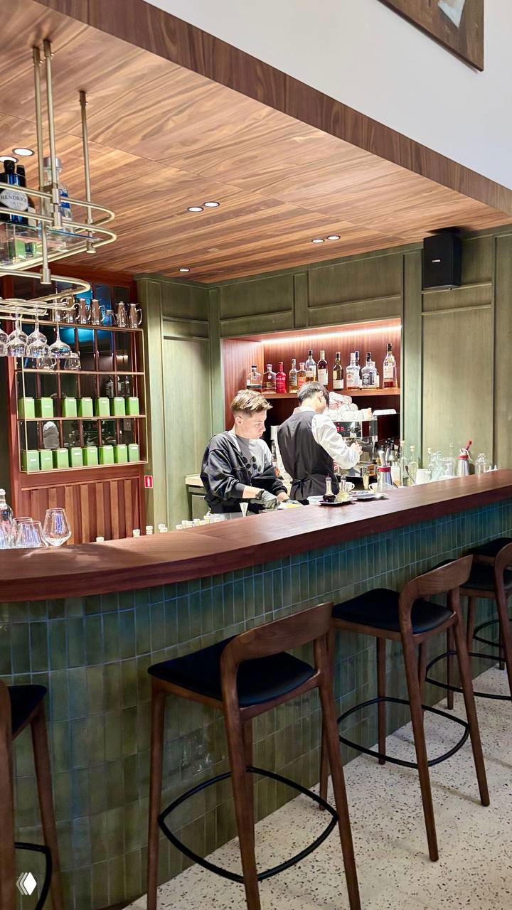 Барная стойка The Mob: деревянная столешница, табуреты, бармены и полки с бутылками на фоне зелёной плитки.