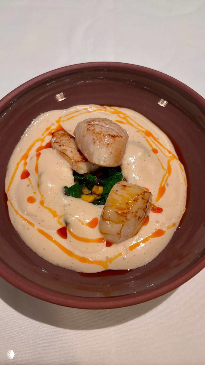 Две крупные обжаренные гребешки, поданные на воздушной пенке; под пенкой видна кенийская фасоль, кукуруза и шпинат.