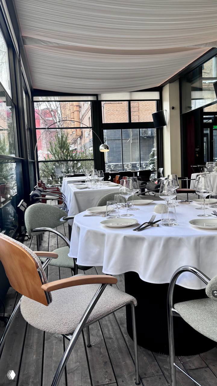 Интерьер ресторана с большими окнами, сервированными столами и белыми скатертями; ощущение светлого и спокойного зала.