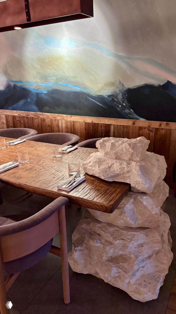 Уютный интерьер ресторана: большой деревянный общий стол, декоративные белые камни как элемент декора и живописная настенная роспись в горах.