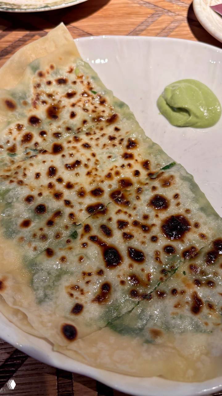 Тонкая обжаренная лепешка (кутаб) с золотистыми прожаренными пятнами и порцией зеленого соуса на белой тарелке — крупный план блюда.