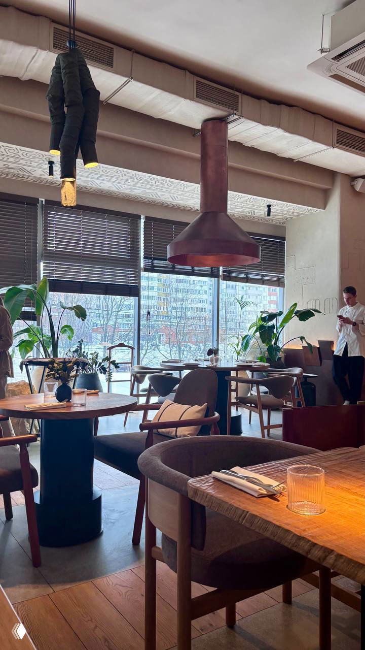 Зона посадки внутри ресторана: столы у больших окон, дизайнерские светильники и медный вытяжной зонтик над открытой зоной кухни, видно город за стеклом.