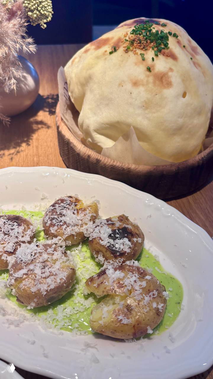 Пышная хлебная шапка в деревянной корзине и тарелка с запеченным молодым картофелем, посыпанным тертым сыром, вид сверху на десктопе.