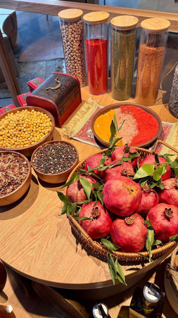 Деревянный стол с корзиной гранатов и разноцветными специями в банках — экспозиция продуктов и пряностей, создающая атмосферу восточного рынка в ресторане.