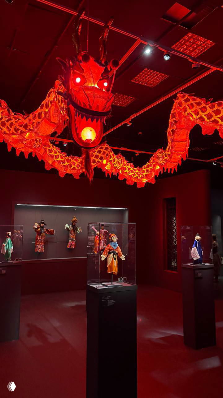 Большая красная драконья инсталляция, подвешенная в зале над витринами с театральными куклами и масками, придающая экспозиции праздничный и театральный вид.