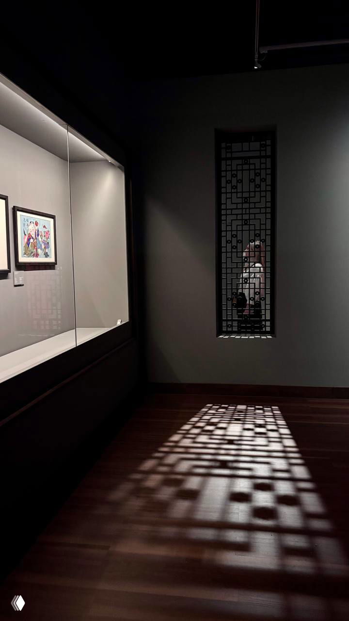 Тёмный зал музея с витриной и декоративной резной решёткой; свет, проникающий через окно, отбрасывает узор на деревянный пол и создаёт атмосферу покоя.