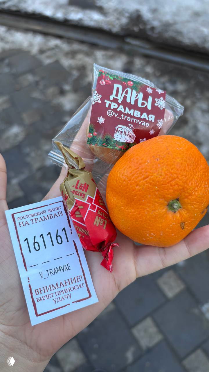 Рука держит мандарин, трамвайный билет с номером и маленькие конфеты/подарки — угощения, розданные участникам спектакля в новогоднем настроении.