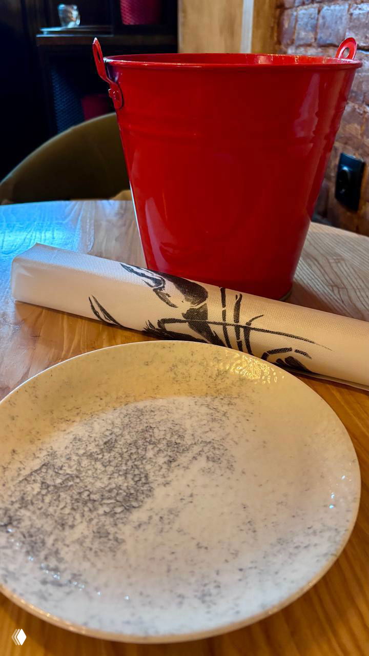 Красное металлическое ведёрко для очисток на столе, рядом скатерть/салфетка и тарелка — столовая сервировка в заведении.