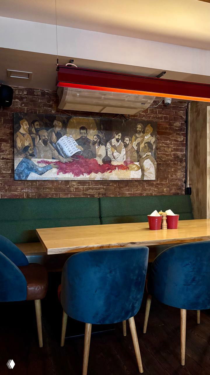 Интерьер ресторана: деревянный стол, синие стулья и зелёный диван на фоне кирпичной стены с настенной картиной «Тайная вечеря».
