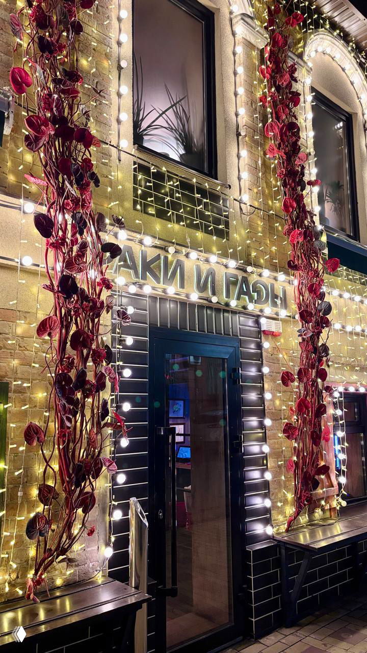 Нарядный фасад ресторана ночью: вывеска «РАКИ И ГАДЫ» окружена гирляндой и вертикальными красными декорациями у входа.