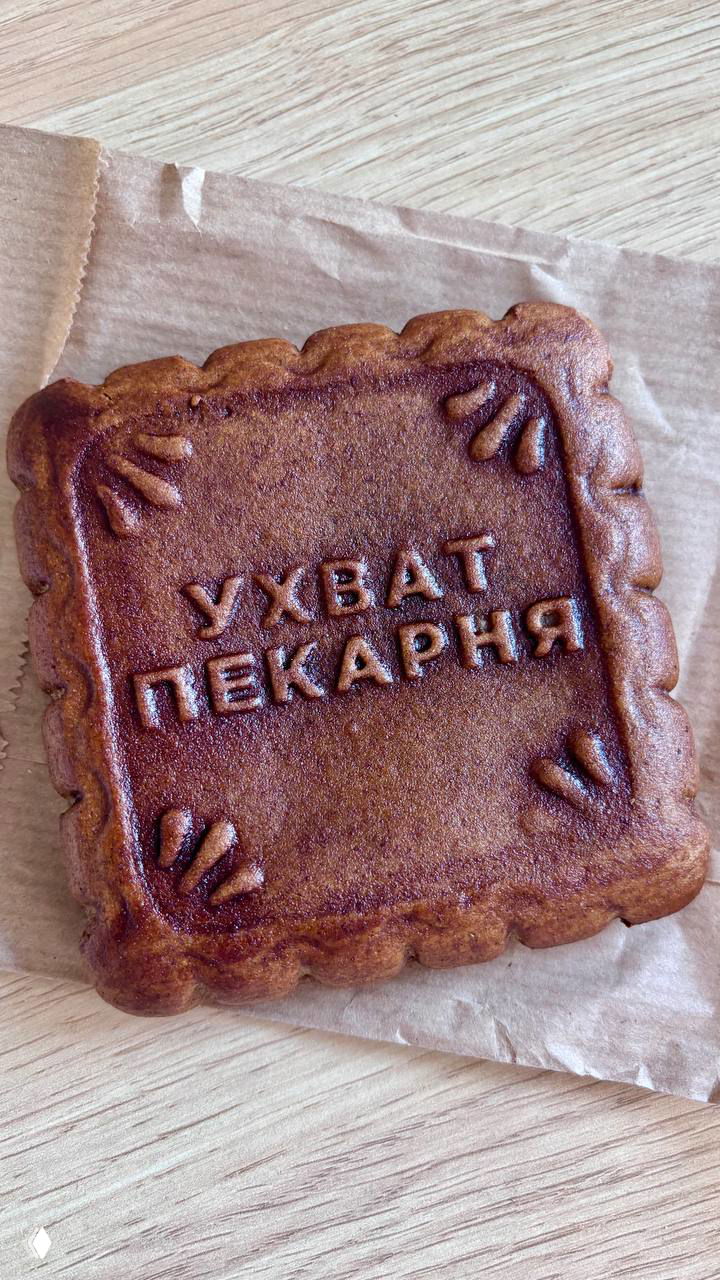Пряник прямоугольной формы с тиснением «УХВАТ ПЕКАРНЯ» на листе пергамента, крупный план узора и румяной корочки.