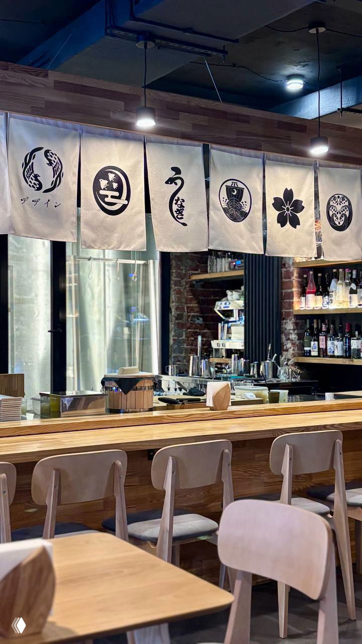 Барная стойка ресторана Shokunin с японскими ширмами (норен), деревянными стульями и открытой кухней, вид спереди на рабочую зону.