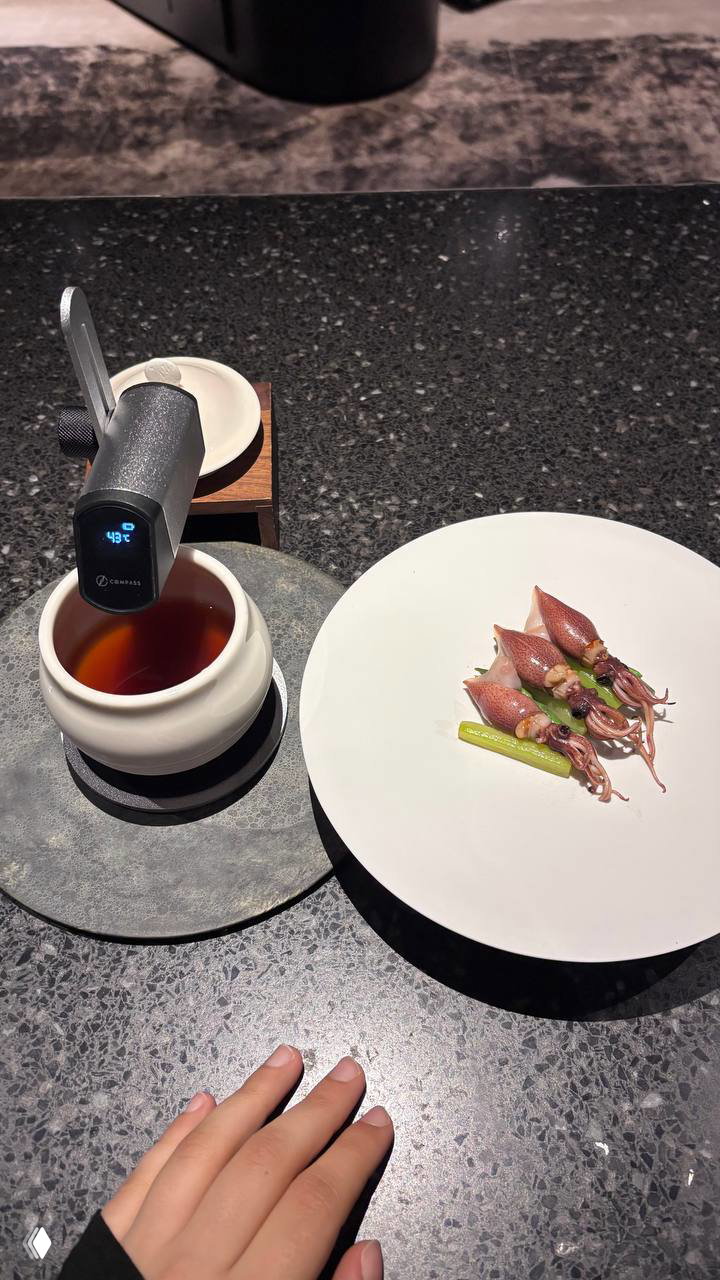 Подача: белая чашка с тёмным напитком рядом с тарелкой мини-кальмаров — контрастная сервировка на тёмной столешнице, видна рука посетителя.