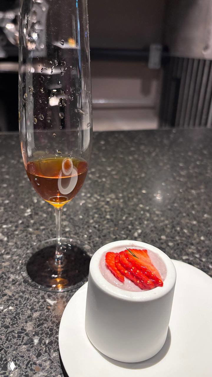 Фужер с янтарной жидкостью рядом с керамической чашей, сверху которой клубничный мусс и слайсы свежей клубники — десертная подача к кофе.