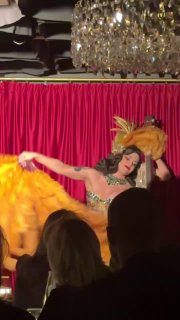 Артистка бурлеска в ярком жёлтом костюме с огромными перьями на сцене бара Louis; передний план — силуэты зрителей, люстра и занавес создают атмосферу.