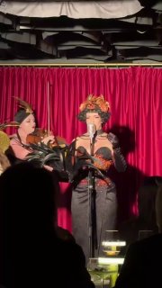 Две исполнительницы бурлеска в театральных костюмах и головных уборах выступают на фоне красной занавеси в баре Louis; одна держит микрофон, публика в тени.