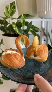 Эклеры-лебеди на тарелке крупным планом, видна золотистая корочка, крем и пальцы держащего блюдо, на заднем плане растения.