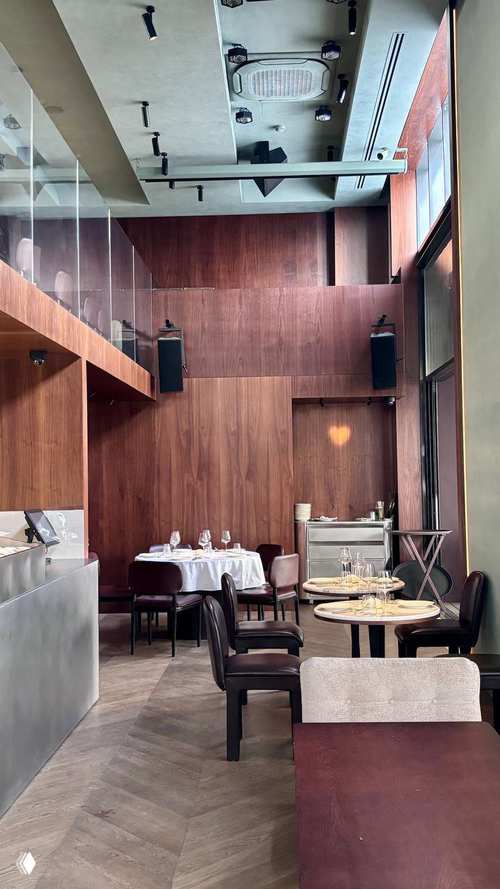 Высокий внутренний дворик ресторана с деревянной отделкой, парой столиков у окна и высоким потолком — современная элегантность.