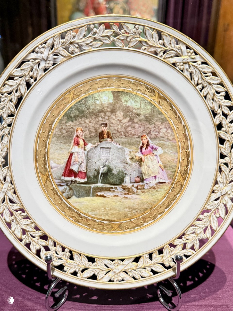 Декоративная тарелка с миниатюрной сценой: две девушки и юноша у родника, богато украшенная рамка — тарелка из коллекции Юсуповых, демонстрируемая в витрине.