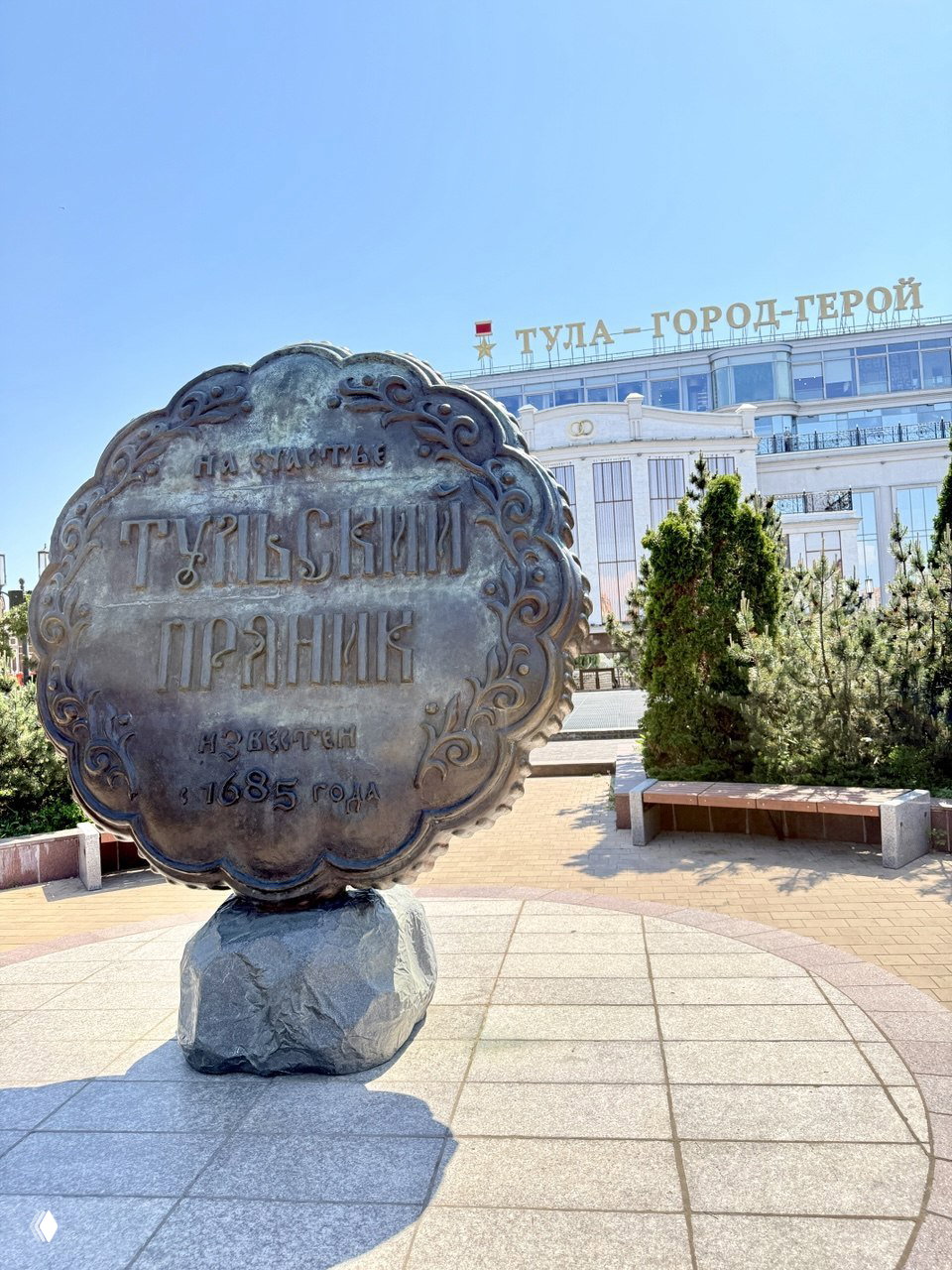 Памятник в виде большого пряника с надписью «Тульский пряник» на фоне центральной площади Тулы и здания с надписью «Тула — город-герой»