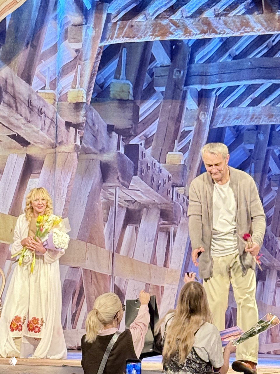 Финал спектакля: актёры принимают цветы и аплодисменты, видно декорации чердака и зрителей, поднимающих букеты и телефоны.