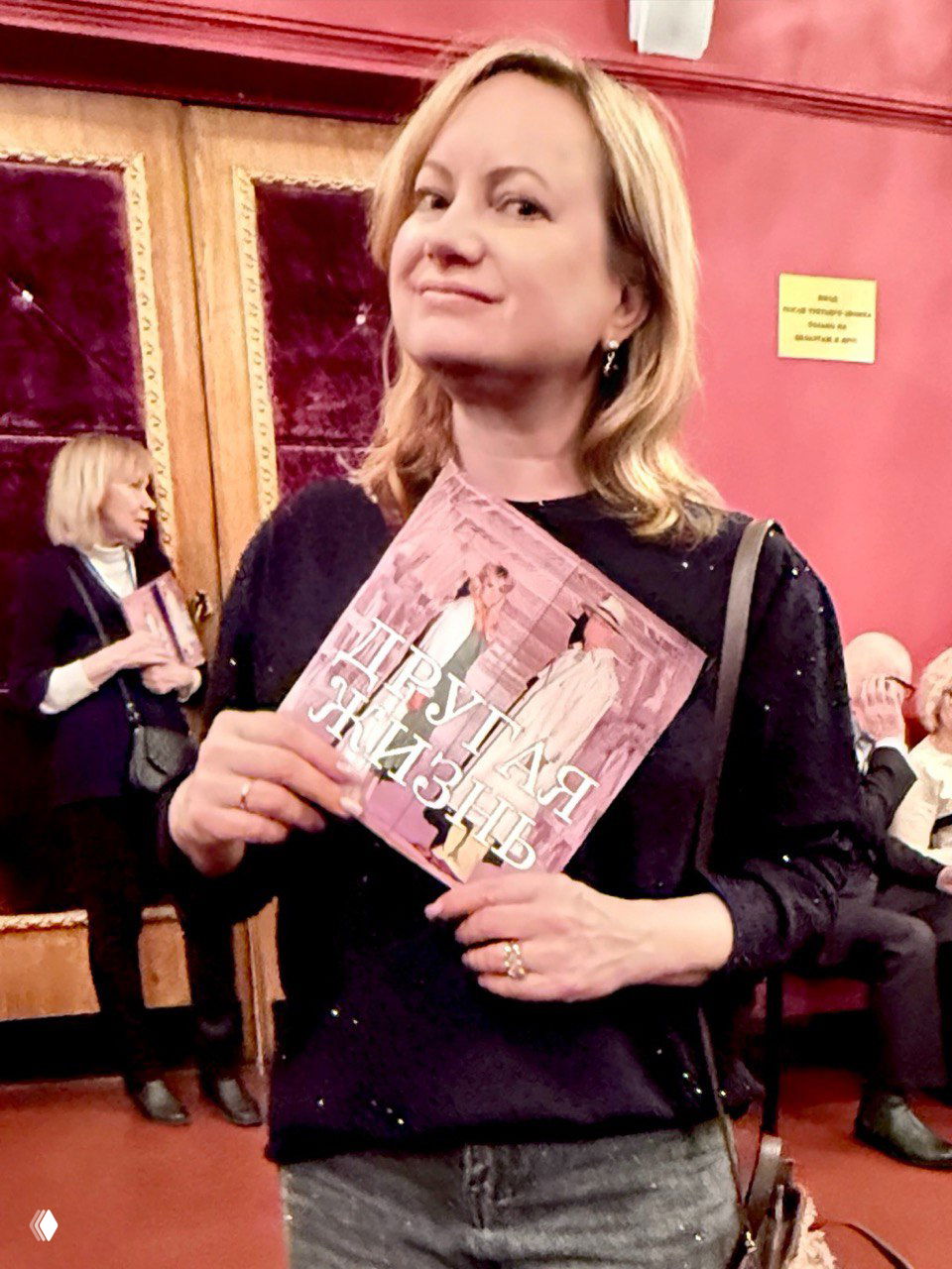 Автор поста в фойе театра держит программу спектакля «Другая жизнь» на фоне дверей и зрительного зала, лицо удовлетворённое после показа.