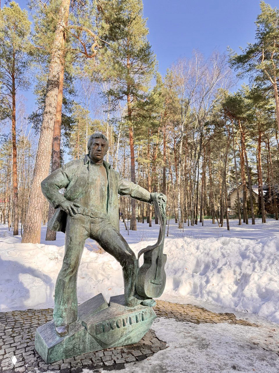 Бронзовый памятник с гитарой в дубнинском парке: фигура музыканта на постаменте среди сосен и снежного покрова в солнечный день.