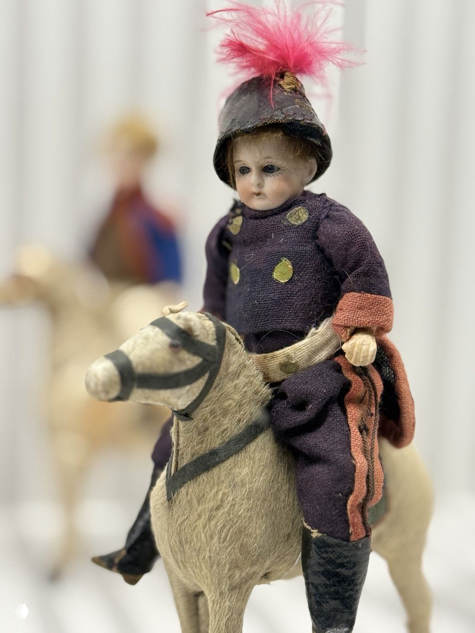 Миниатюрная кукла‑солдатик в тёмном костюме с розовым помпоном на шляпе, сидящая верхом на игрушечной лошадке — витринный экспонат с аккуратной отделкой костюма.