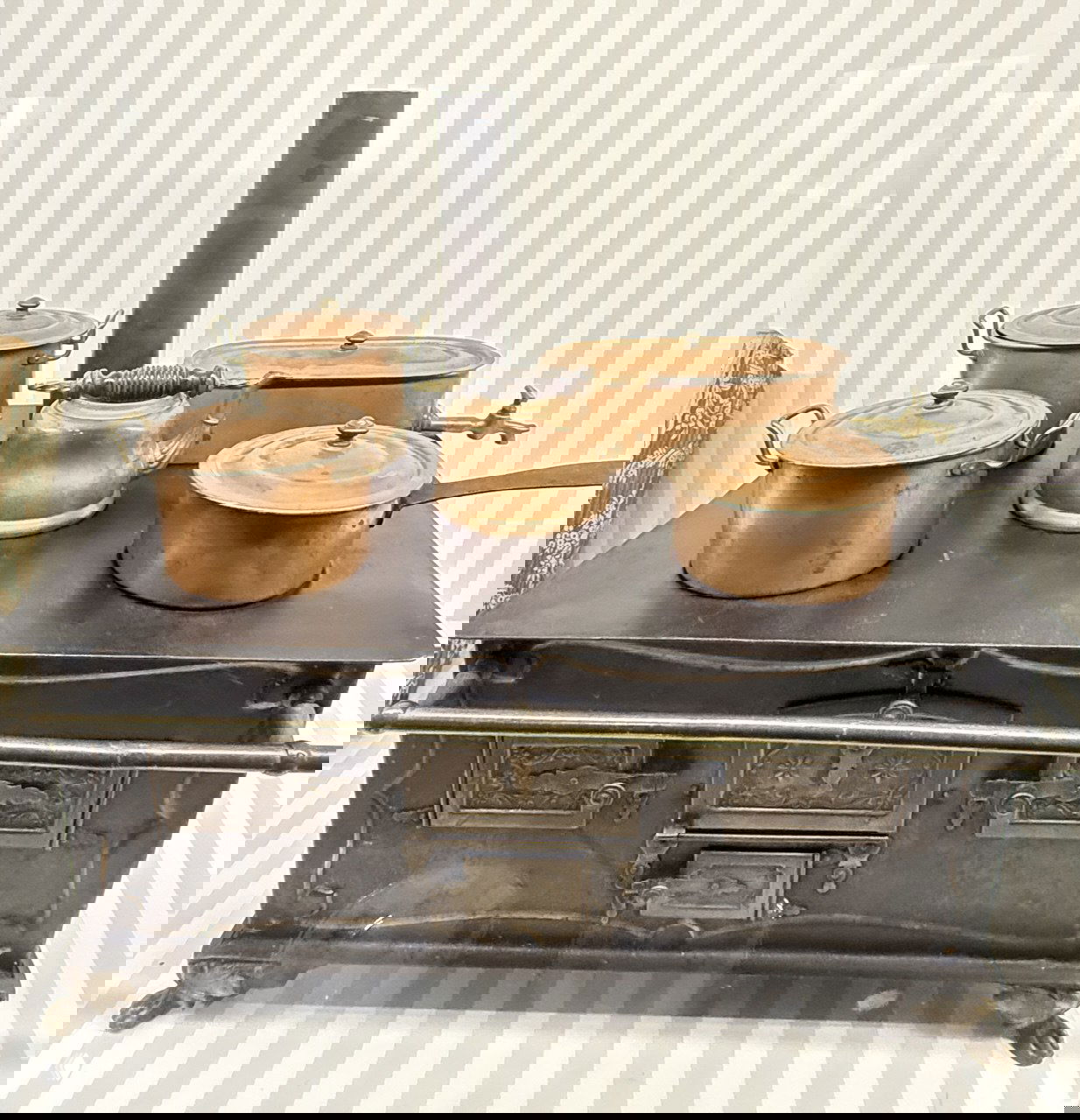 Миниатюрная чугунная плита с медными кастрюлями и чайником — уменьшенная кухонная утварь, демонстрирующая быт в кукольном доме на выставке.