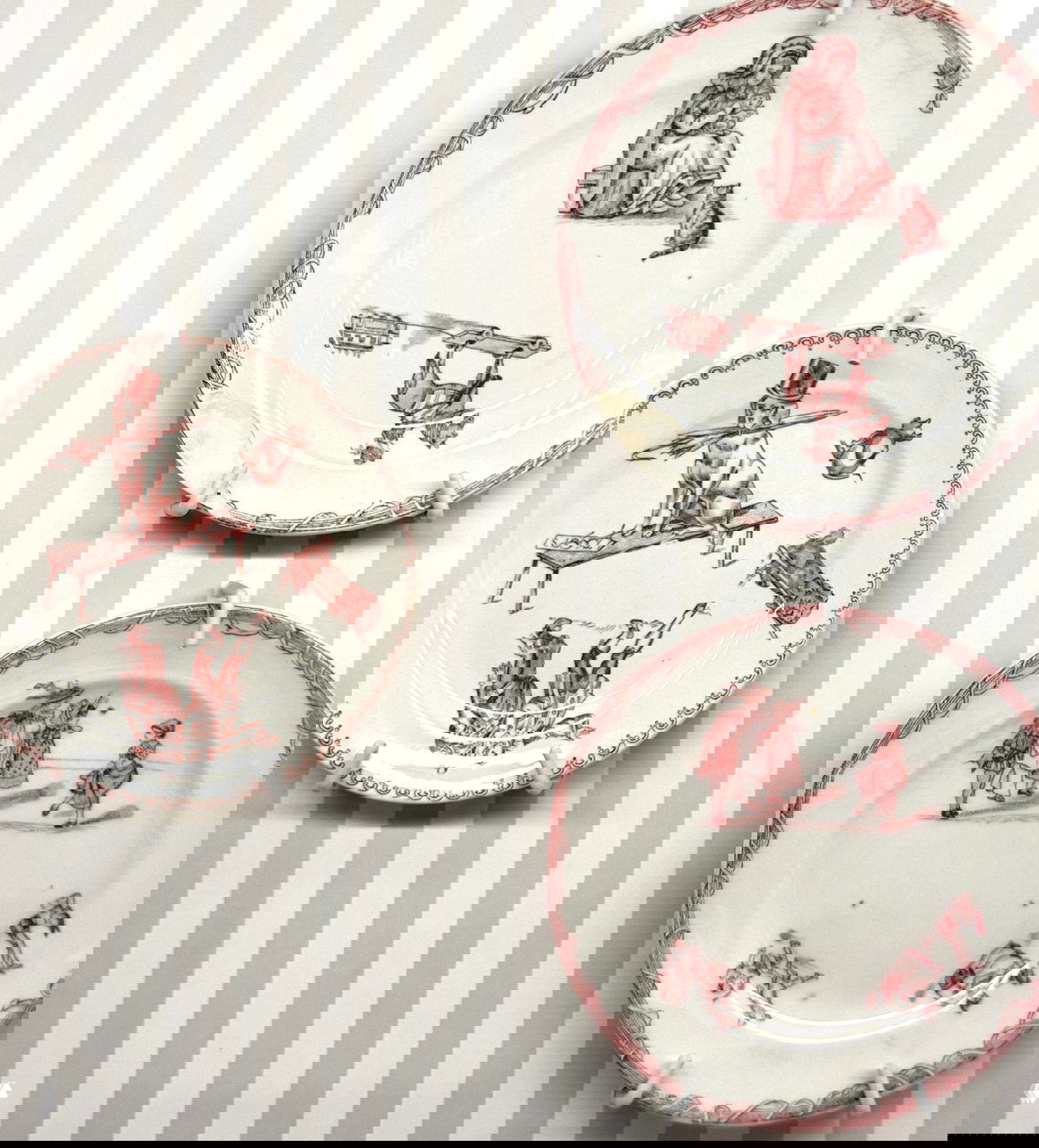 Керамические тарелки с красными жанровыми сценами на фоне полосатой витрины — примеры сервизов ручной работы, показанные рядом с куклами.