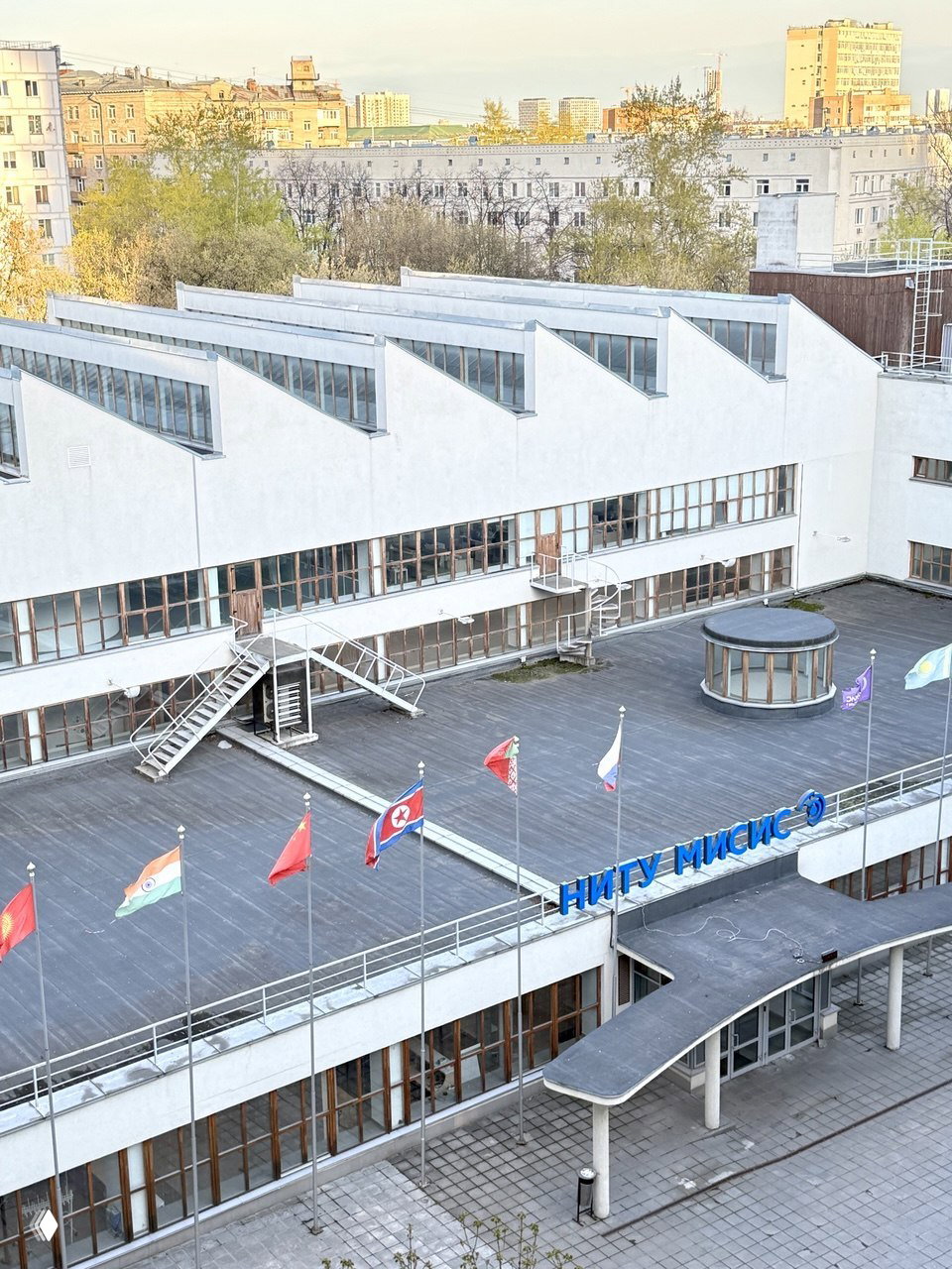 Крыша с зубчатыми окнами и широкой террасой корпуса, на которой установлены флаги разных стран и виден вход в НИТУ МИСиС.