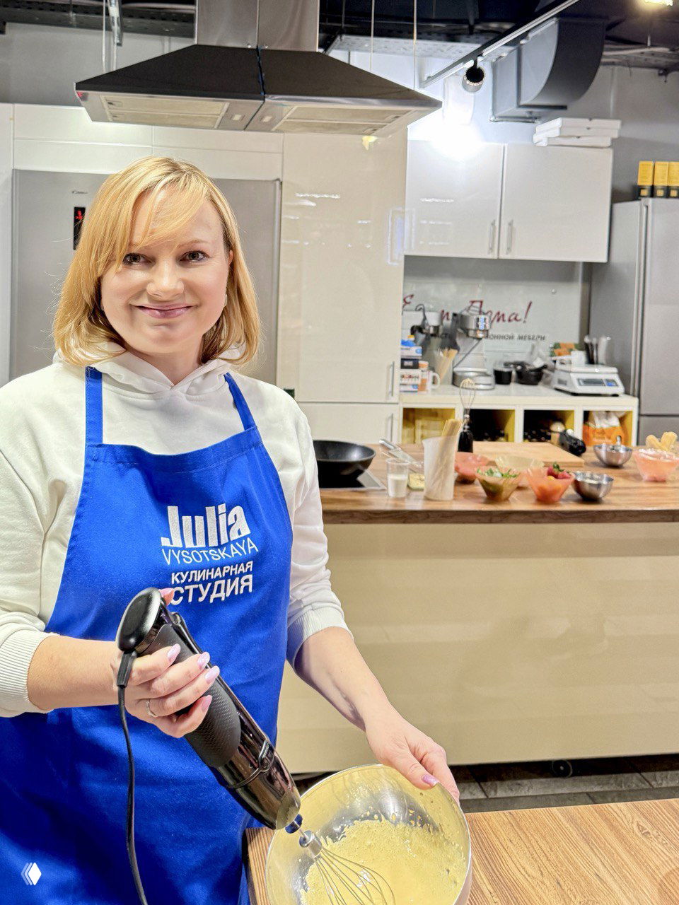 Автор в фартуке с надписью Julia Vysotskaya держит погружной блендер и миску с кремом в светлой современной кулинарной студии