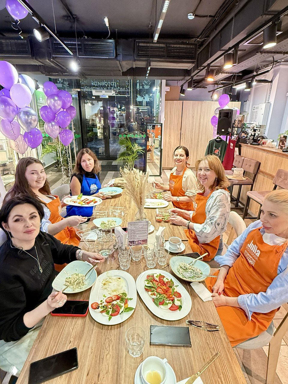 Группа участниц за длинным общим столом в студии: все в фартуках пробуют приготовленные блюда, на заднем фоне витрины и украшения
