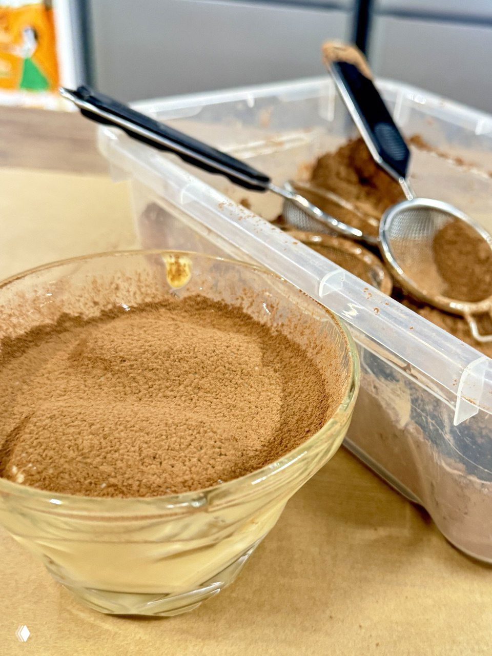 Крупный план чашки с тирамису, щедро посыпанной какао; рядом контейнер с какао и кухонные сита — готовый десерт мастер-класса