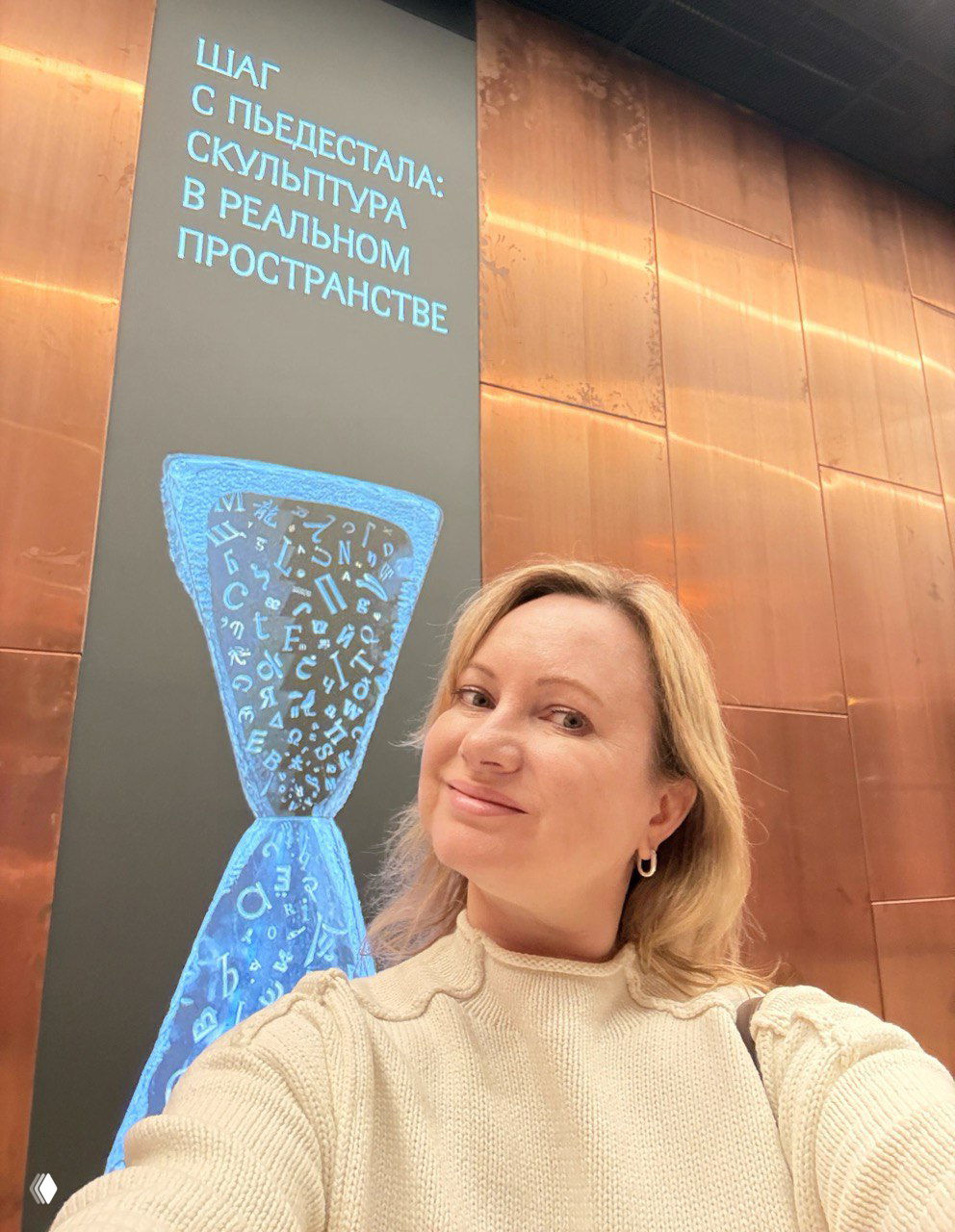 Селфи женщины в холле ЗИЛАРТА: автор улыбается на фоне вертикального баннера выставки «Шаг с пьедестала», медные стены и интерьер зала.