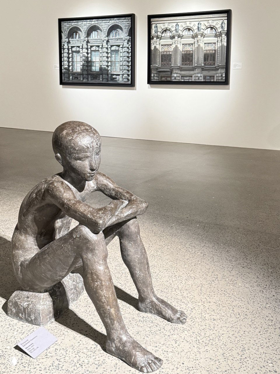 Бронзовая скульптура сидящего мальчика на полу галерейного зала на фоне парных рамных работ и ровного пола, фотоснимок экспоната крупным планом.