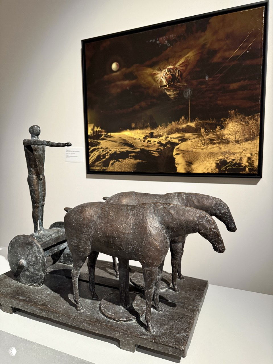 Бронзовая композиция: три лошади на платформе и фигура на колеснице на переднем плане, за ней висит живописное полотно с тёмным пейзажем.