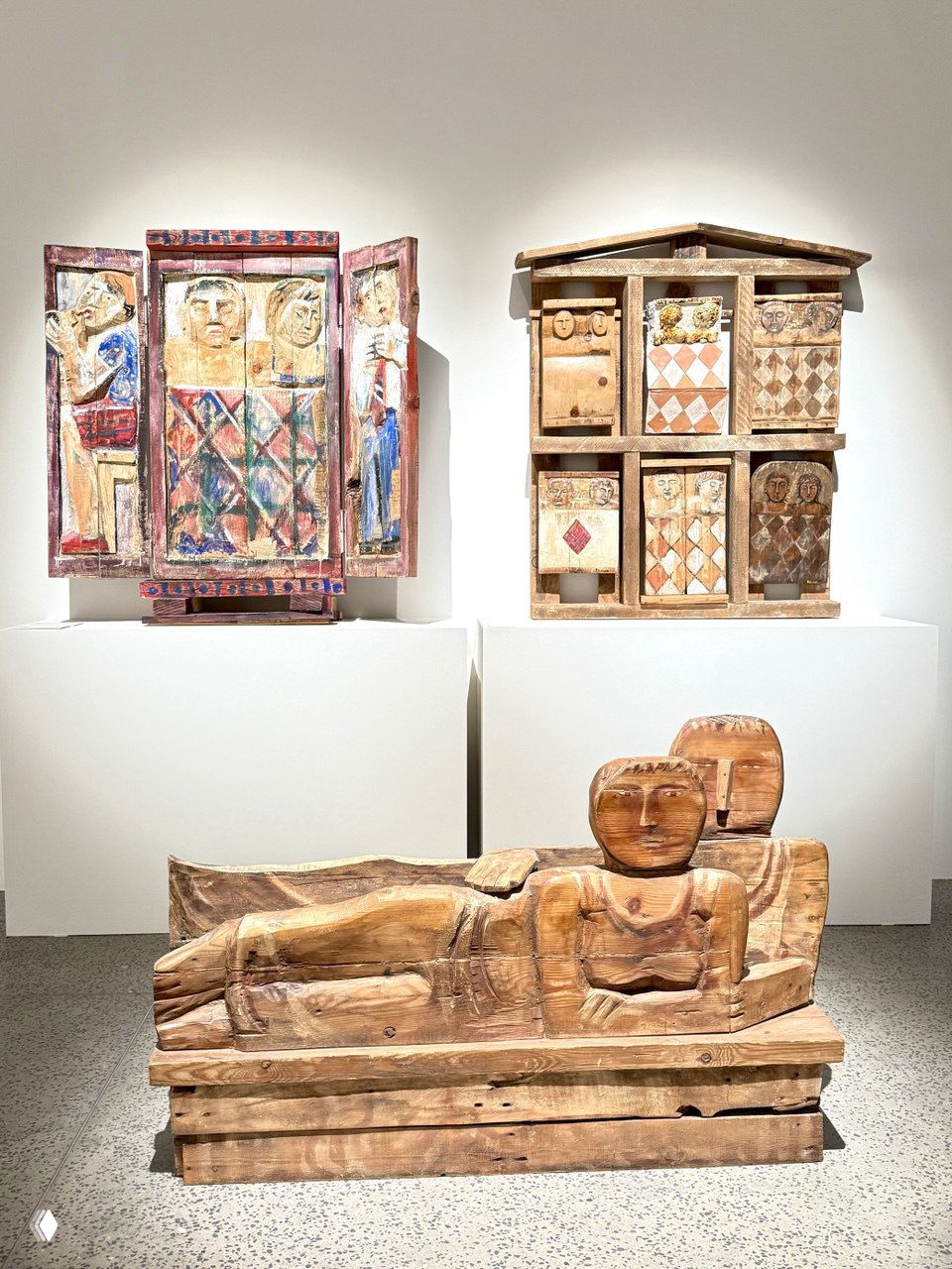 Группа деревянных резных скульптур и расписных панелей на белых постаментах: народная пластика и натурные объекты в экспозиции музея.