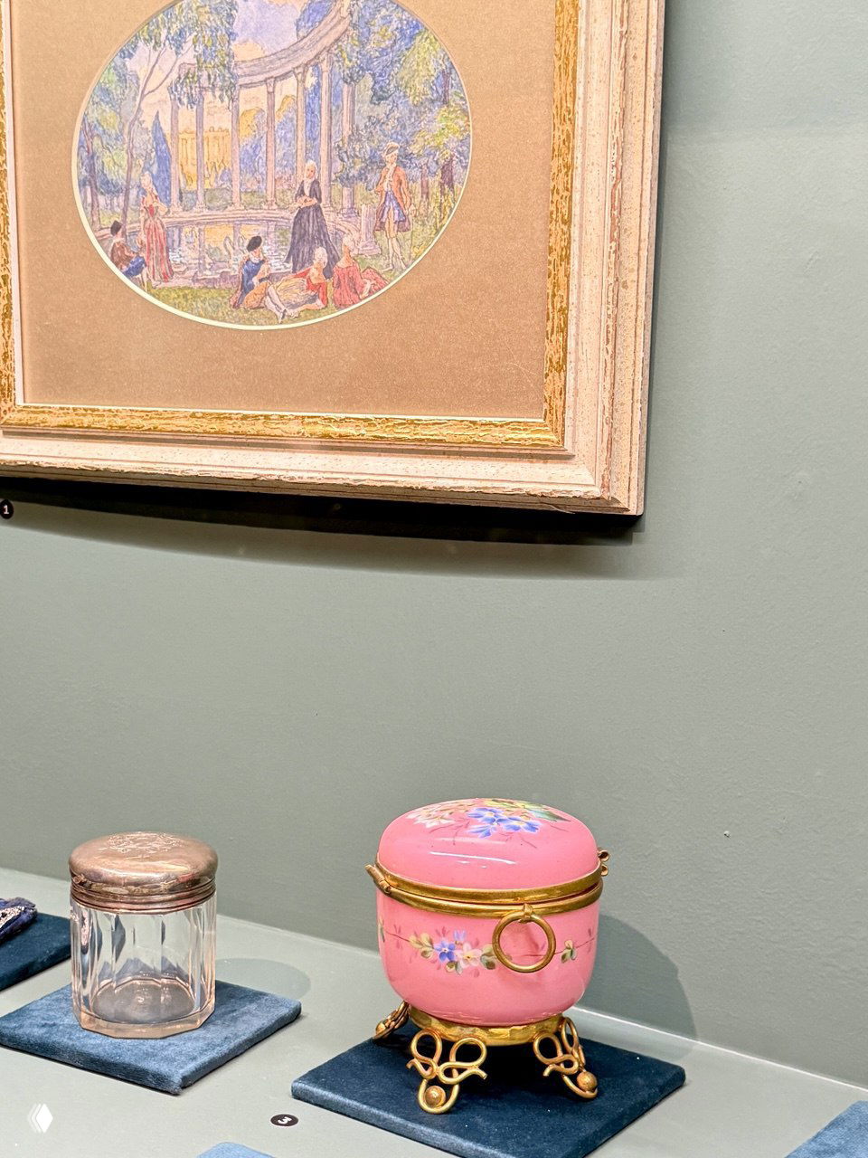Миниатюрные музейные предметы на подставках — стеклянная баночка и розовая шкатулка с позолоченным основанием на фоне картины в раме.