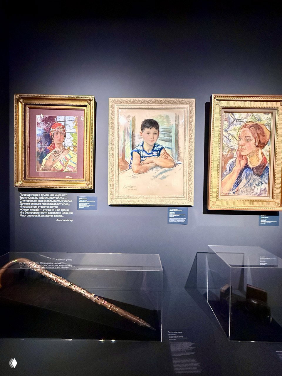 Три картины в рамках на тёмной стене и витрины с музейными предметами на переднем плане — классическая выставочная витрина.