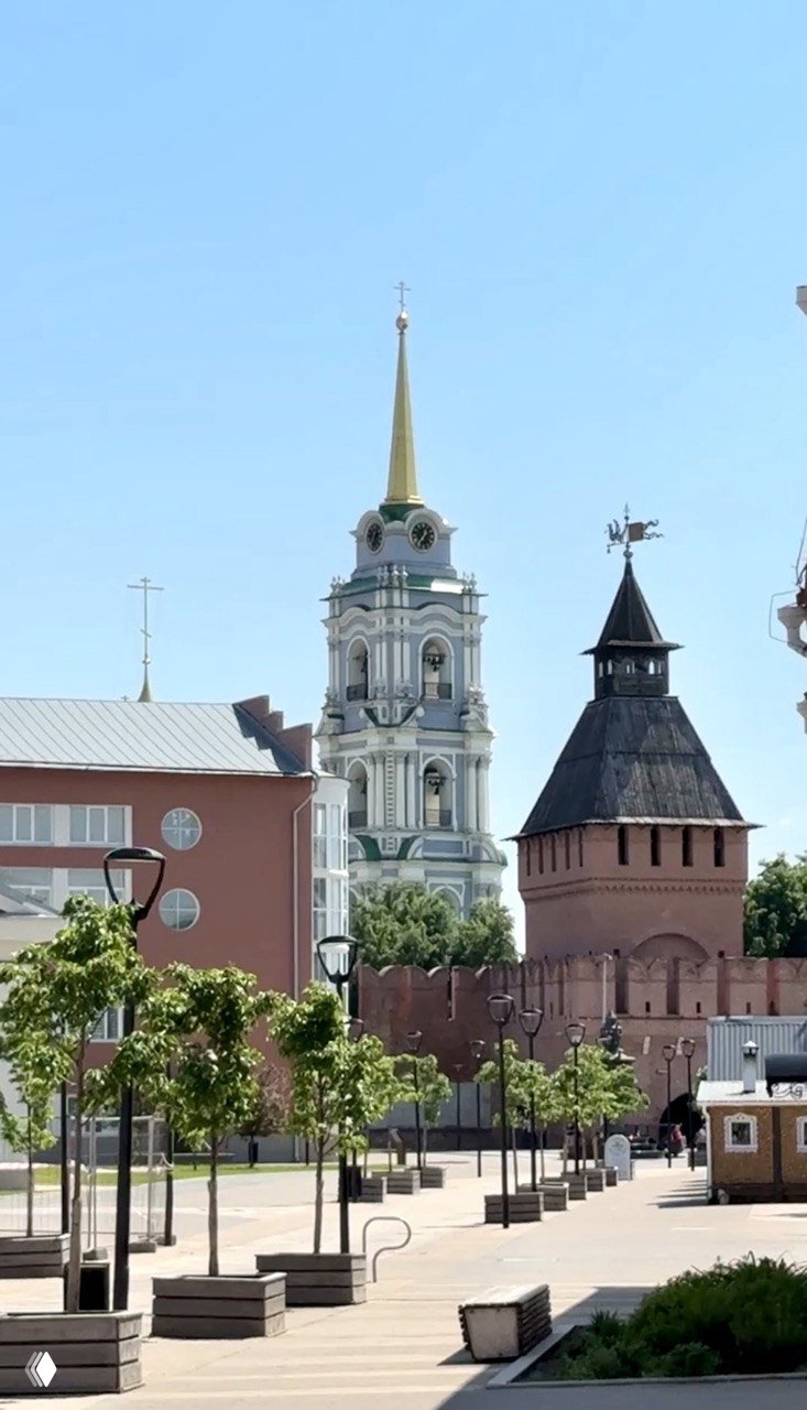 Панорамный вид на колокольню и одну из башен кремля со спокойным голубым небом и частично видимыми крышами окружающих зданий.