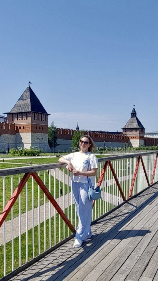 Женщина позирует на мосту возле кремля; в кадре видны башни и крепостные стены на заднем плане, солнечный и зелёный городской пейзаж.