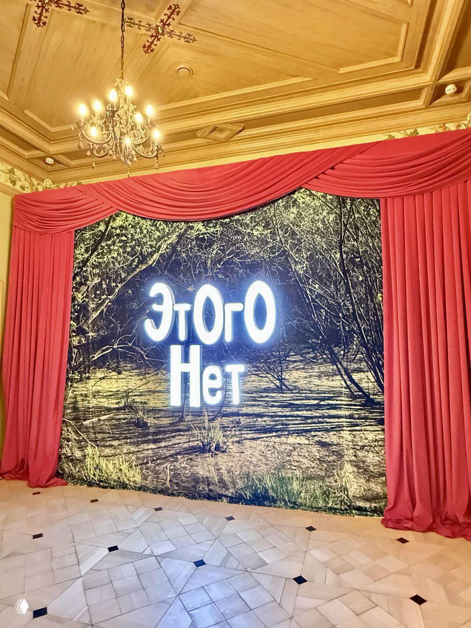 Сцена с широким полотном и неоновым словом «Этого Нет» в светлом зале, оформленная красными занавесями и люстрой над выставочным пространством.