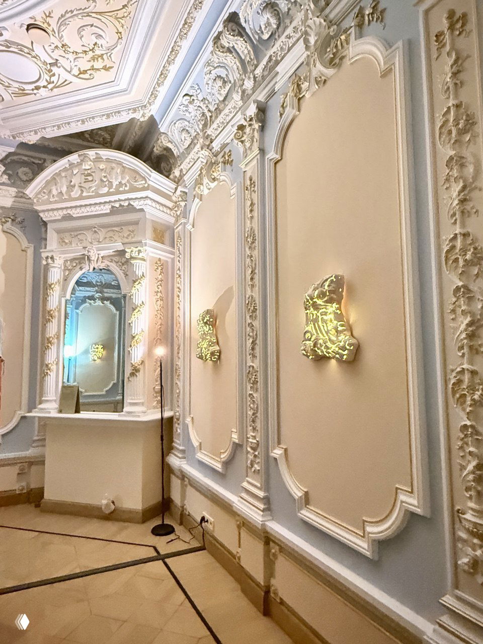 Интерьер в пастельных тонах с барочными стеновыми панелями и небольшими светящимися объектами-барельефами, расположенными по периметру зала.