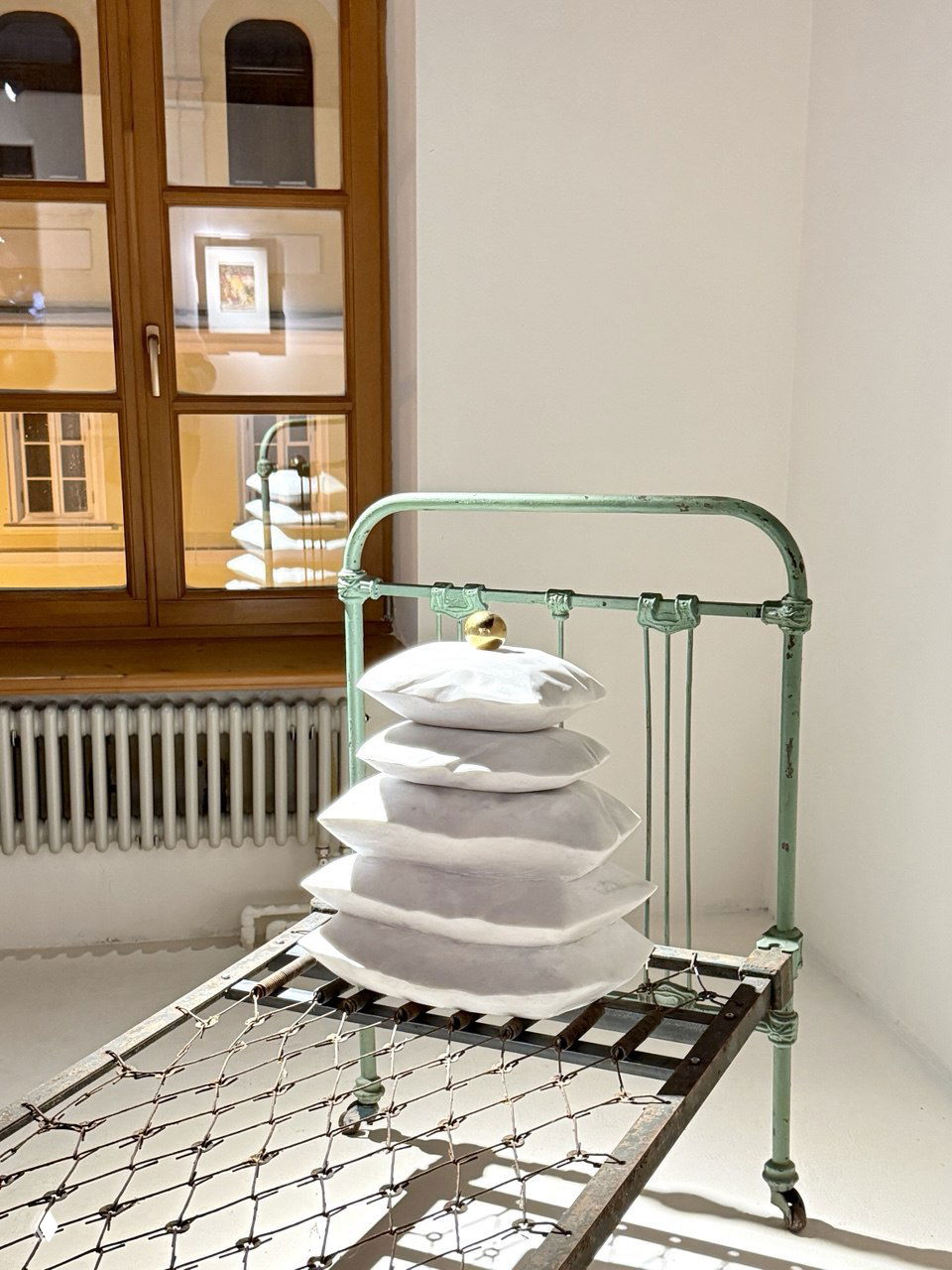 Старый металлический каркас кровати салатового цвета с сетчатым основанием и стопкой белых подушек как художественный объект в минималистичной белой комнате.