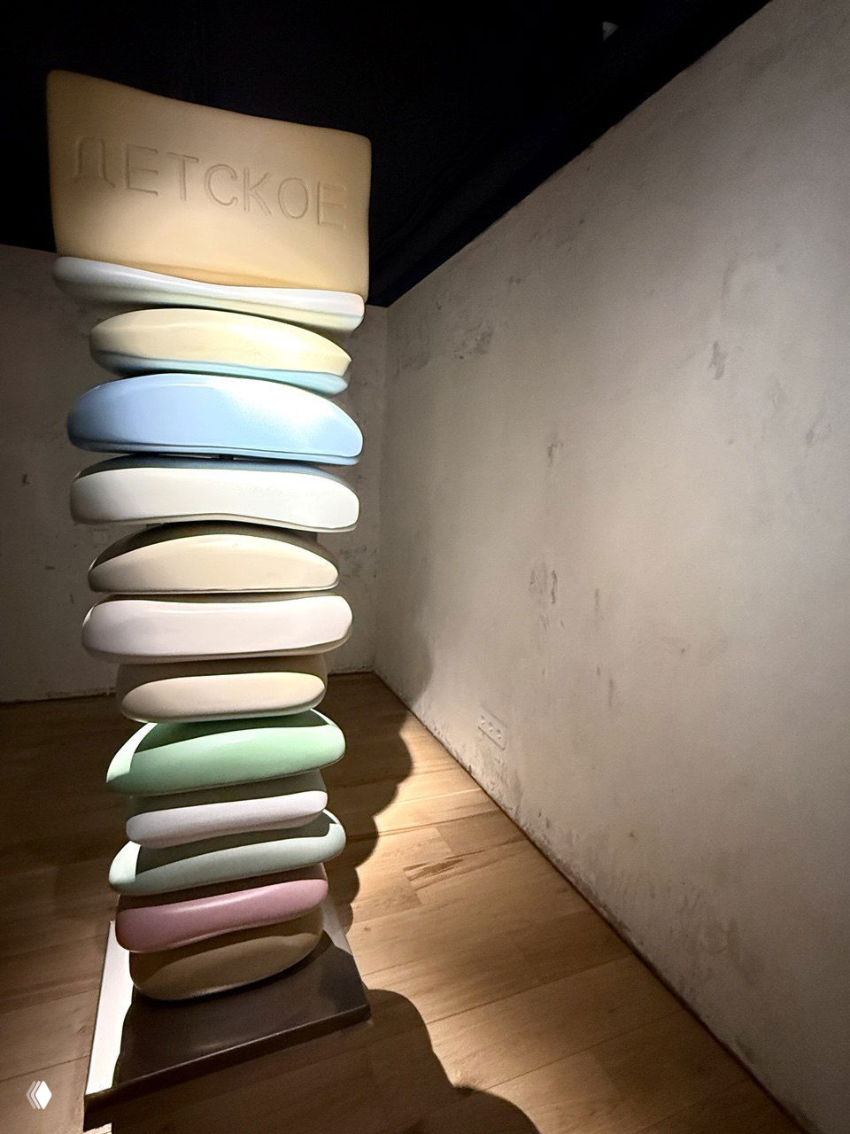 Палевая колонна из нескольких пастельных объёмных форм, напоминающих мыло или подушки, с верхней табличкой «ДЕТСКОЕ» — предметная скульптура в углу зала.